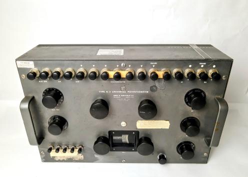 Potentiometer is comprised of a large metal box with several knobs on the top panel. These have labels such as: 'Aux EMF', 'Aux Pot', 'Galvanometer', 'range', 'SC', 'fine,' 'coarse, ' and 'GA sens.' An analog dial is labeled "Volt" and has a display of numbers that can be manipulated by turning the knobs. There are also two handles on both ends of the box. Instrument label includes Leeds & Northrup catalogue #7553 and serial number #1527443
